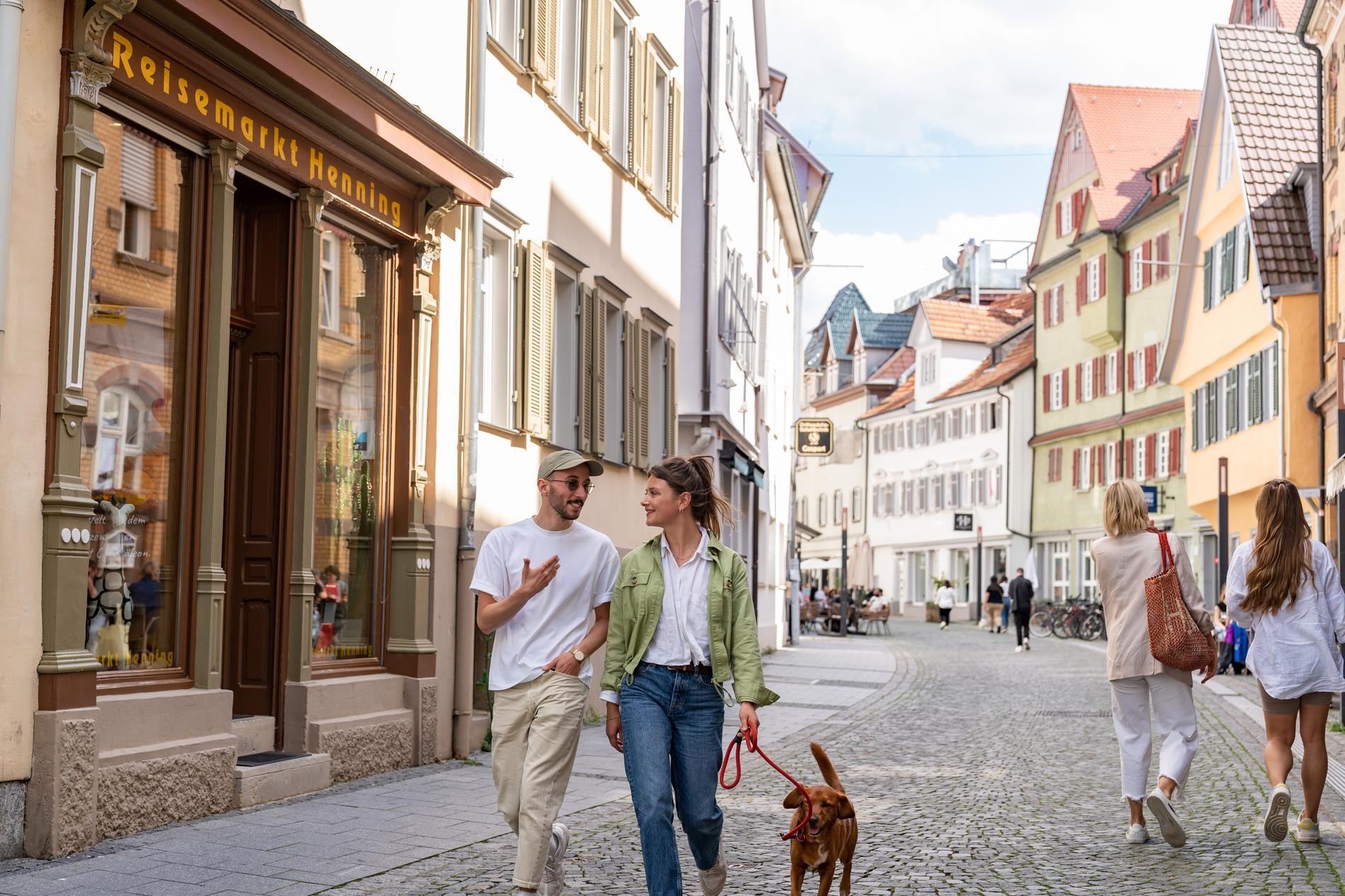 Eine Frau und ein Mann mit einem Hund spazieren durch eine Fußgängerzone. Rechts und links sind bunte Häuser mit bunten Fensterläden zu sehen.