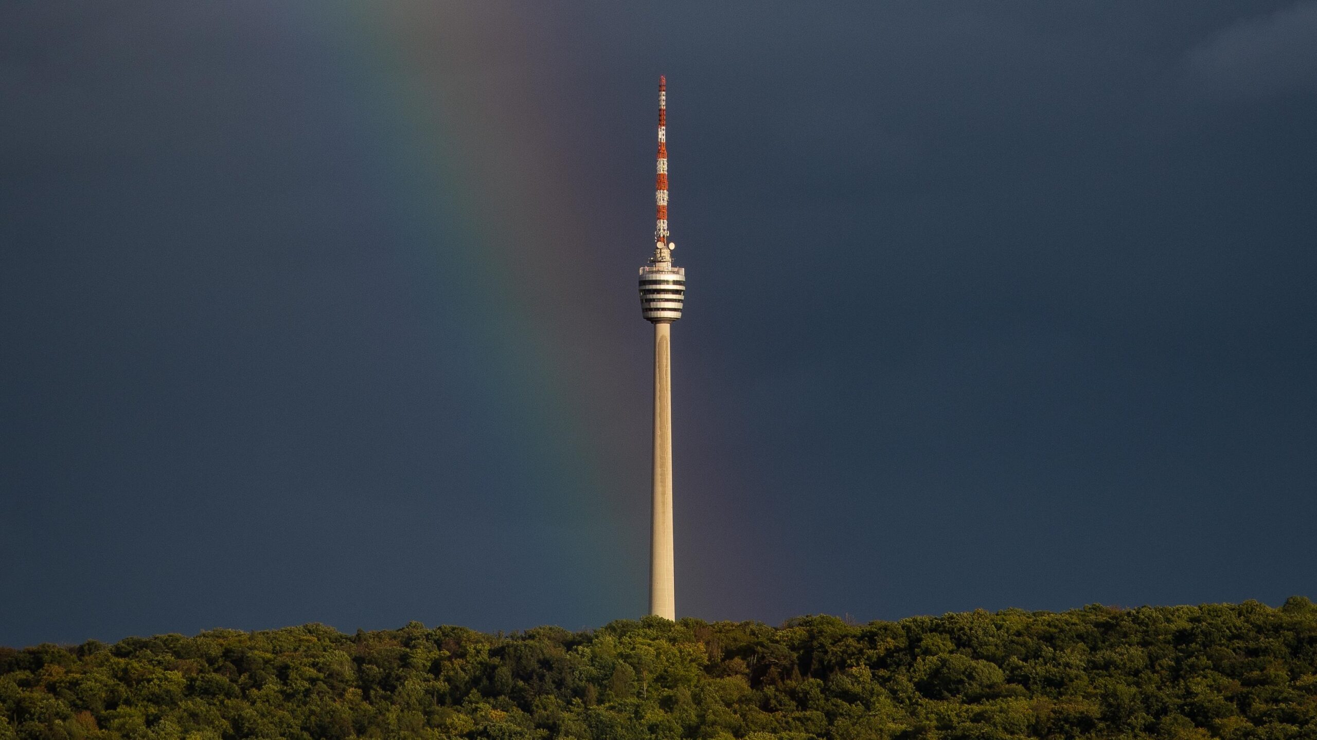 Der Stuttgarter Fernsehturm ragt über grünem Wald, ein Regenbogen erscheint schwach vor dunklem Himmel. Ein Kontrast aus Natur und Technik.
