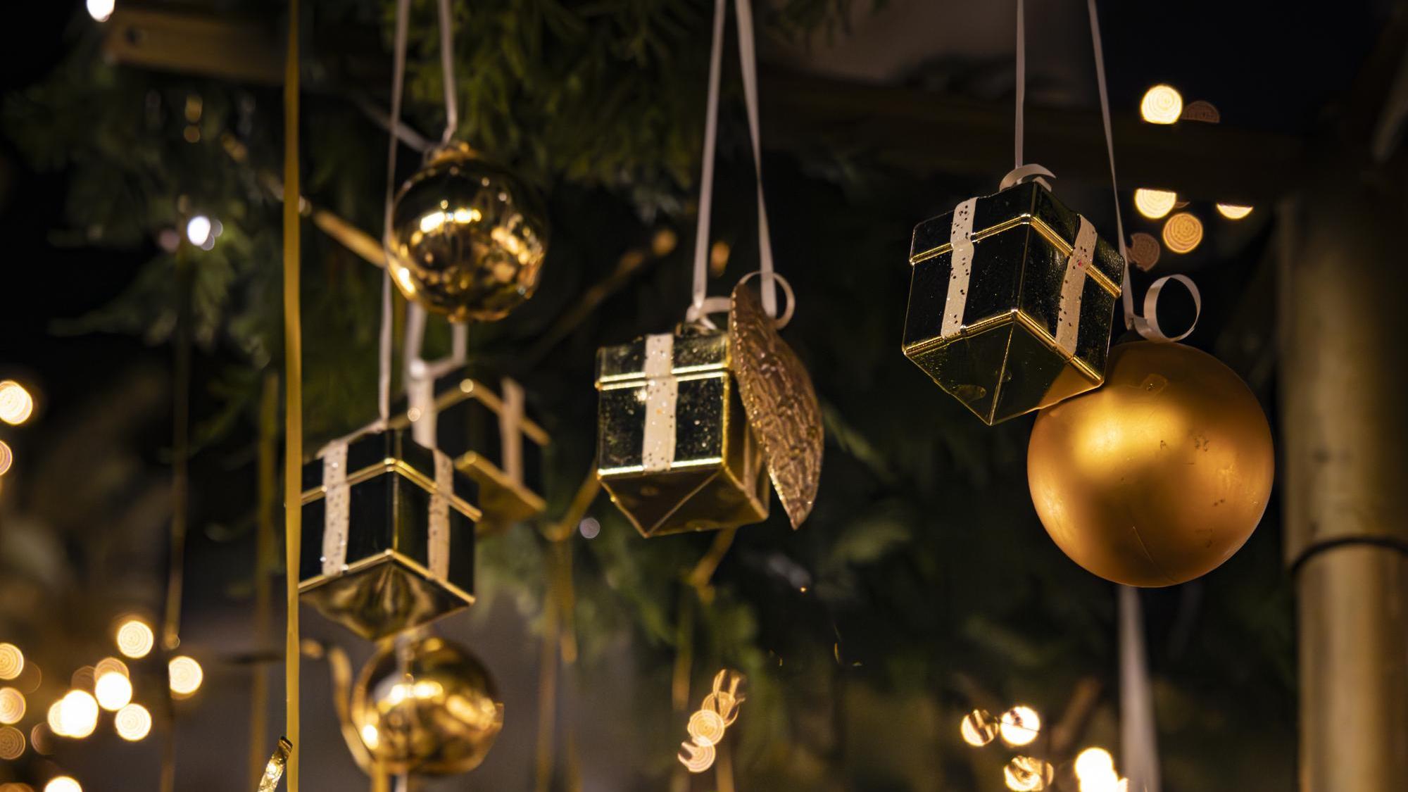 Goldene Kugeln und kleine Geschenkboxen hängen als Dekoration an einem Weihnachtsbaum. Im Hintergrund leuchten warme Lichter unscharf.