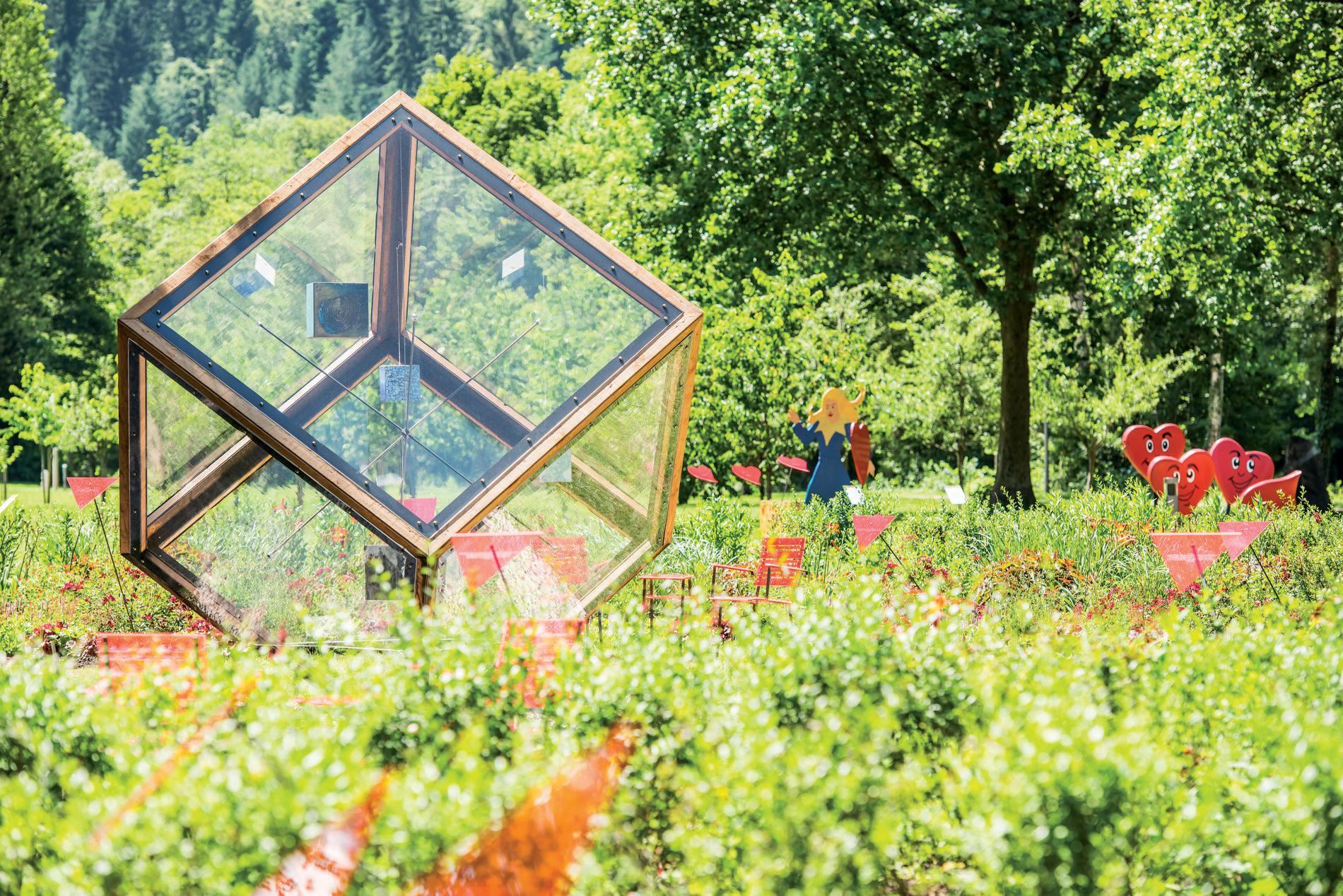 Moderner Glaswürfel als Kunstobjekt in einem bunten Garten. Dahinter grüne Bäume, rote Herzfiguren und bunte Deko.