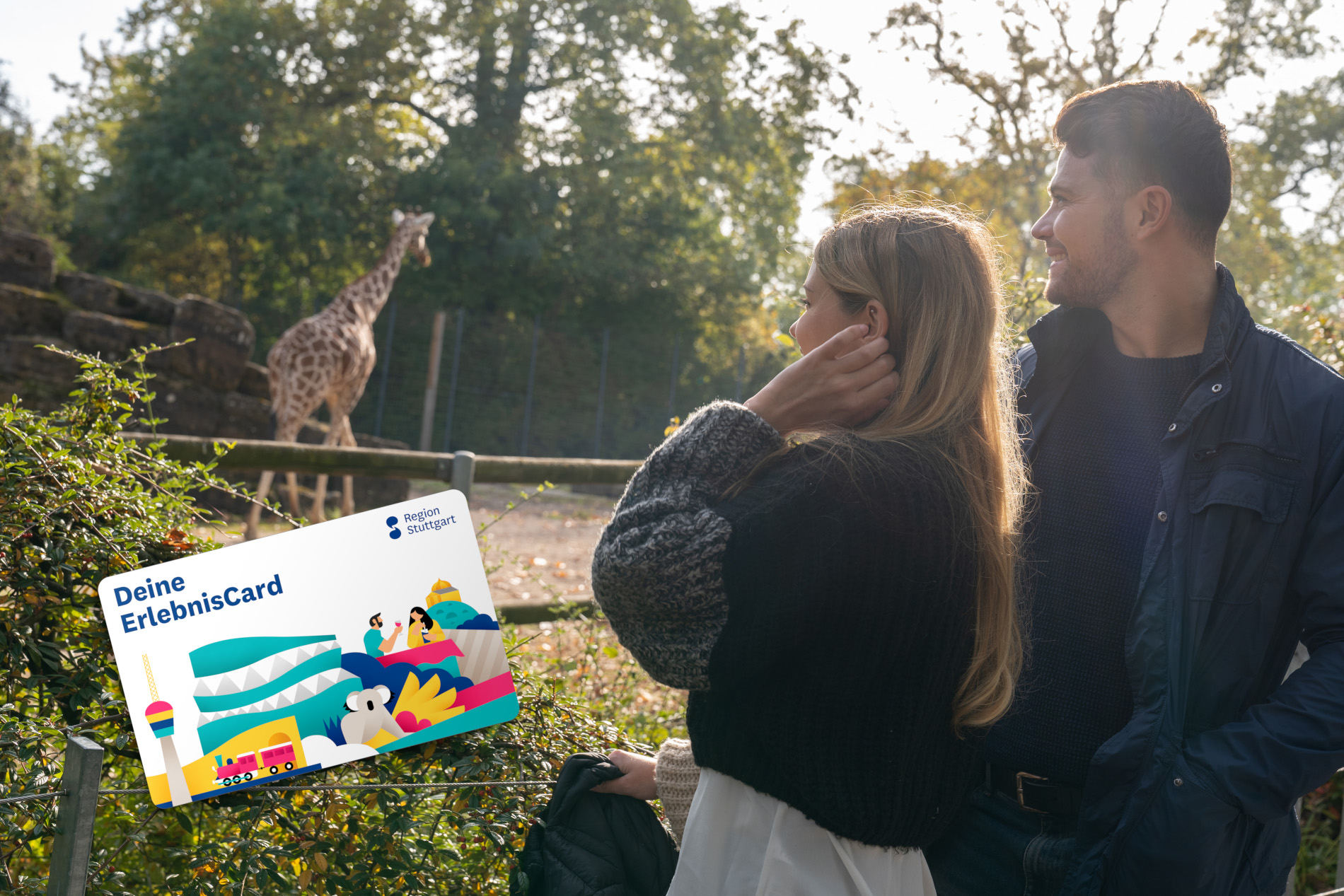 Ein Mann und eine Frau schauen in Richtung einer Giraffe im Hintergrund. Sie sehen glücklich aus. Auf dem Bild eingefügt ist eine Grafik der ErlebnisCard der Region Stuttgart.