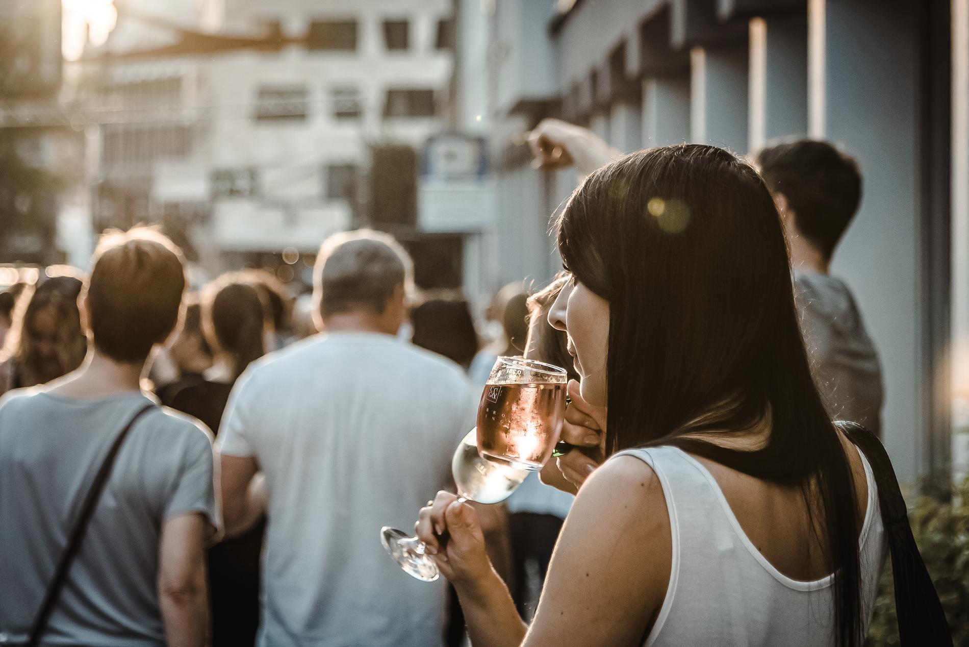 Der Fokus liegt auf einer Frau, die auf einer Straße steht und ein Glas Wein bei Sonnenuntergang trinkt. Im Hintergrund sind weitere Menschen zu erkennen.